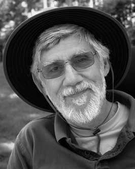 Image Description: Chris Palames is seated outdoors in his wheelchair in a peaceful rural setting. He wears a dark sun hat, glasses, and outdoor attire, with a full gray mustache and beard.